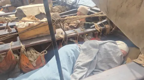 Gward An Aod - Brioc People wrapped in woollen blankets sleeping across the boat between benches, with ropes and sails around them.