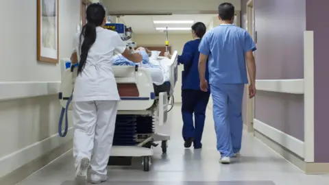 AFP via Getty Images NHS staff in blue and white uniforms wheel a patient trolley down a corridor, viewed from behind