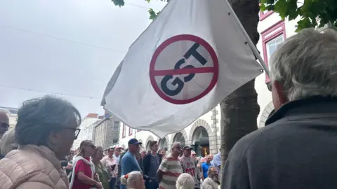 A large flag which says GST in the middle with a red strikethrough going through it. Behind it are people standing some holding anti-GST placards. 