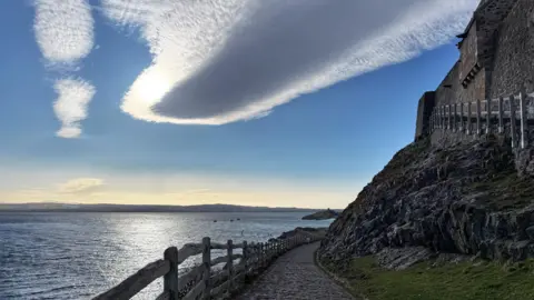 Amelia A stone path with a rough wooden fence looking out on to the sea and to a small island in the distance. It is flanked by an old building built into a sloping rock. There are a few clouds in the blue sky. 