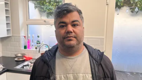 Mehdi Mozadeh - a man with short, greying hair looks at the camera. He is wearing a grey jumper and a black zip-up jacket. He is standing in a white kitchen, with some dishes in the sink and cleaning products on the window sill.