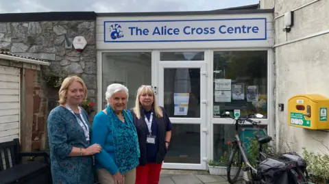 Three members of the staff of the centre stood in front of the entrance. It's a concrete and strong building with large glass doors. 
The three woman in the from are stood together looking at the camera. Two of them have blonde hair and one of them has grey hair. A bicycle is parked by the main entrance. 