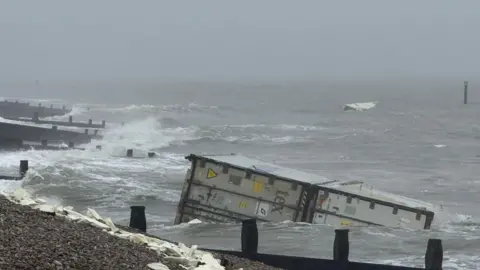 A shipping container washing up on a stony beach.