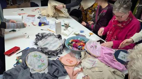 Emergence Restock A close-up of a work table with several items of clothing being embroidered on and a dish of many skeins of different coloured wool. People working on the textiles can be partially seen in the background, looking very busy and concentrating hard.