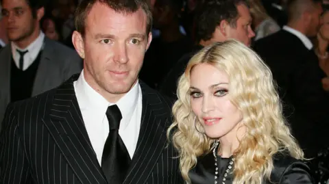 Getty Images Guy Ritchie has dark hair and is wearing a pin stripe suit over a white shirt and black tie. Madonna has blonde wavy air and is wearing a black outfit. They are pictured at a film event.