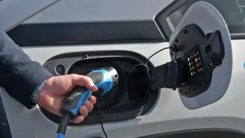 A hand is holding a blue EV charger into the side of a white car. You can see they are wearing a silver watch and dark grey blazer.