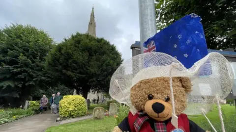 A teddy bear dressed in a white fancy hat and tartan bow tie with a British and Australian flag with the churchyard in the background