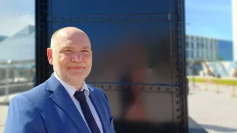 Peter Richards is smiling. He has a navy suit on with a white shirt and a dark navy tie. Behind him is a large steel sculpture. 
