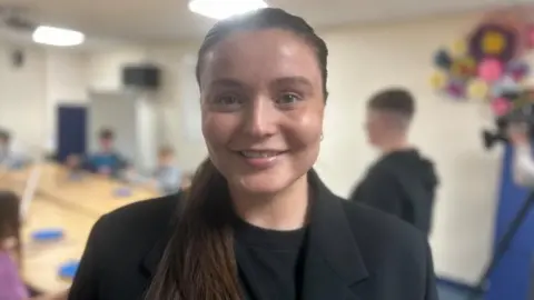 Zara is wearing a black suit jacket and black t-shirt. She has long brown hair tied back in a pony tail style. She is smiling at the camera.