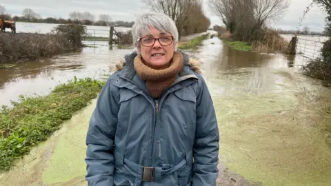 Paula, a middle-aged woman with grey hair and red glasses. She is wearing a brown roll-neck top and a blue coat, and is standing in front of a flooded landscape