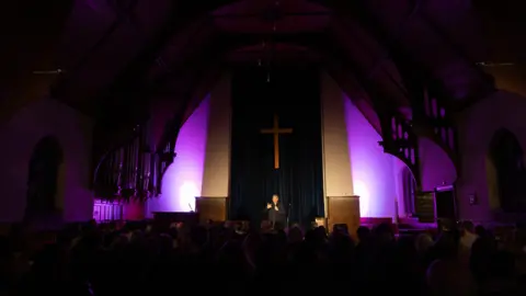 Priya Dabasia A stage inside a church with purple lighting. A man is talking on the stage in front of a crowd.