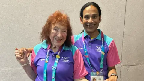 A 96-year-old woman with red slightly wavy shoulder length hair is smiling at the camera. She is wearing a blue, pink and purple short sleeve, collared Commonwealth Games t-shirt. Next to her is a woman who has her arm around her. She is wearing the game top and smiling at the camera. She has black long hair in a ponytail. 