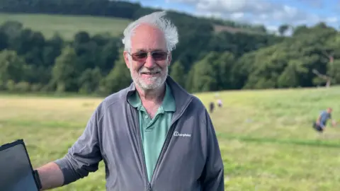 A grey haired man with dark glasses and a beard smiles at the camera. He's wearing a grey jacket with a green shirt beneath. Behind him are people working in a field.