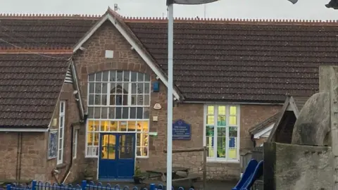 The Old Cleeve School building. There is a blue entrance door and a sign next to it. The building is made of red bricks and has big windows with white frames.