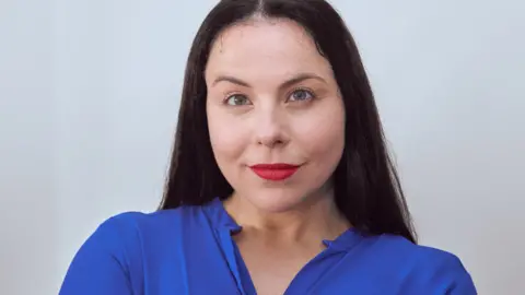 Amy Leibovitz A woman with long dark hair is smiling at the camera. She is standing in front of a white background and is wearing red lipstick and a blue blouse.