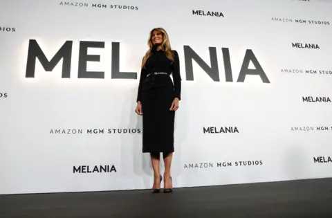 The First Lady of the United States Melania Trump stands smiling at the premiere for her documentary Melania. She is wearing a long sleeved black dress with black high heels and a silver belt, and is standing in front of a banner that has 'Melania' written in large black capital letters. She is standing in the centre, in front of the middle 'A' in Melania. 