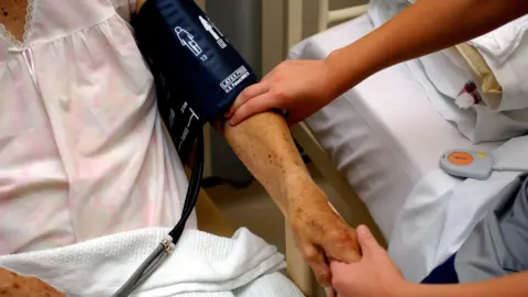 An elderly woman wearing a pink nightdress, getting her blood pressure checked by another person sitting on a bed next to her. The faces of the people cannot be seen in the picture.