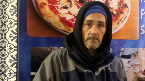 A man stands behind a picture of a pizza at a fast food restaurant. He is wearing a black hoodie and a blue beanie hat.