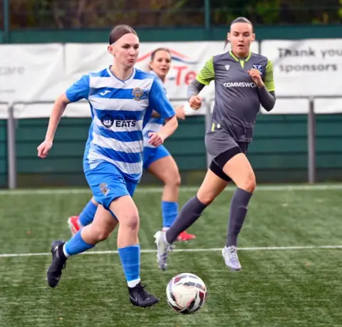 Taylor Coulter Three women playing football. A womanin a blue striped top is dribbling the ball with a player in a different strip nearby 