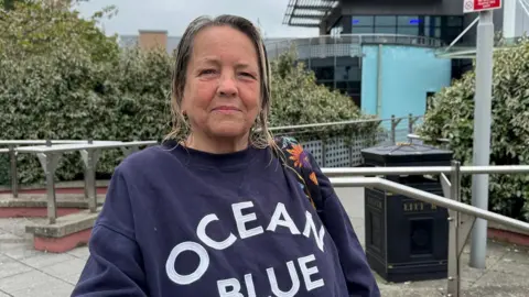 Julia is wearing navy jumper that says ocean blue. She has long highlighted hair which shes wearing half up half won