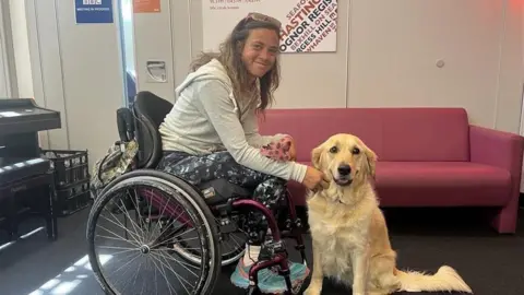 Jazz Turner in an office in her wheelchair with her assistance dog Phoebe, a golden labrador