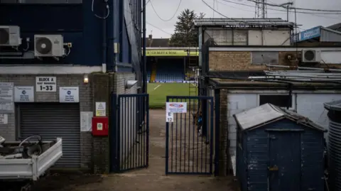 Getty Images A gate behind Southend's Roots Hall stadium, leading to the pitch. There are various small buildings that look run down in the foreground. In the background, the green grass of the pitch and blue seats in a stand are pictured.