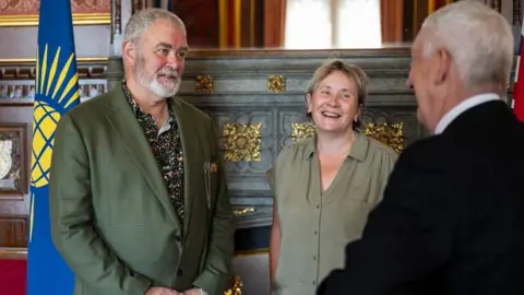 Terry Wiggins, wearing a greenish  blazer and patterned shirt, faces the camera, arsenic  Lindsey Hoyle (seen from behind) talks to him 