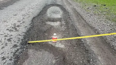 Andrew McQueen A plastic bottle is sticking out of a pothole, with further road damage around it.