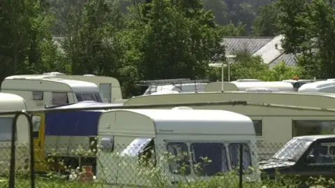 White caravans stationary at a site which is behind a wire fence. 