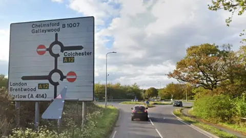 A Google streetview image of Stock Road in Chelmsford, near junction 16 of the A12. There is a large sign to the left indicating two roundabouts and the directions to head to enter the A12. At either side of the road is green shrubbery and grass. The sky is blue with some clouds.