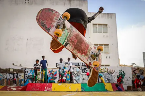 Cem Ozdel / Getty Images A young skateboarder performs a flip in the air while others look on.