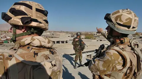 Two British Army soldiers wearing desert fatigues stand close to the camera in an urban landscape in Afghanistan. An Afghan National Army soldier stands, framed by the British troops.
