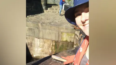 Eve Ingle Eve is wearing an orange life jacket and blue clothing and holding a scrubbing brush against a dark wooden surface near a stone canal lock. The background shows a stone-paved area with moss on the walls and a metal fence, with another individual standing further away.