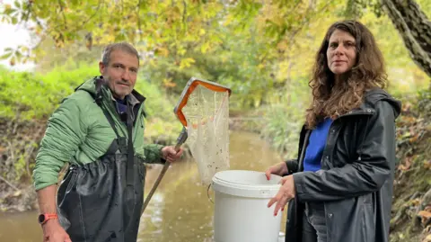 A man and a woman are standing by the River Grom looking at the camera. the man, in waders, in holding a net, while the woman holds a bucket