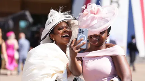 PA Media Two women, both wearing large hats and a white and pink dress respectively, smiling as one on the right takes a selfie at the racecourse.