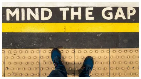 Mind the Gap sign shot taken vertically so you can see the sign on the platform and someone wearing trainers standing behind the yellow line on the bobbled paved section. Above the yellow line there is a Mind the Gap sign painted in white bold capital letters.