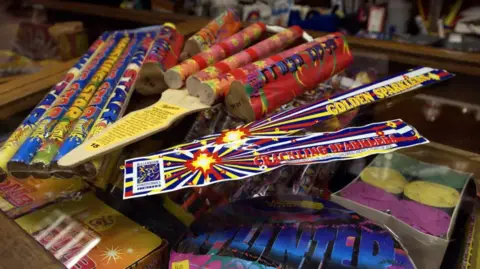 PA Sparklers, rockets and other fireworks sit on the counter of a shop, the products having a very colorful branding. 