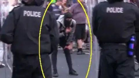 Cleveland Police Still from body worn footage. Two police officers in riot gear have their backs to the camera. A man in black trainers and trousers bends over moving a black balaclava on his face