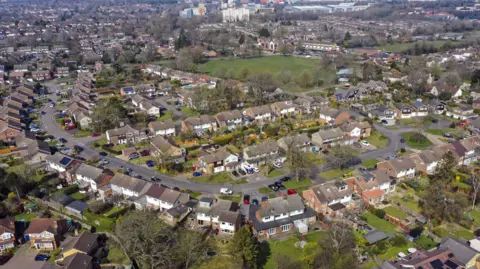 An aerial shot of a low-density suburban neighbourhood with detached and semi-detached houses.
Streets are arranged in curving, cul-de-sac-style layouts, typical of post-war residential planning.
In the background, the landscape transitions into a mix of larger buildings, light industrial units, and open areas.