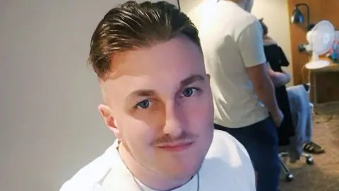 A man with short brown hair looks up at the camera. He wears a white t-shirt and a silver chain