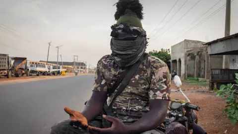 A man wearing a camouflage-patterned top, his face covered with scarves, sits on a motorbike, a gun in his lap and his hands open in front of him, palms up