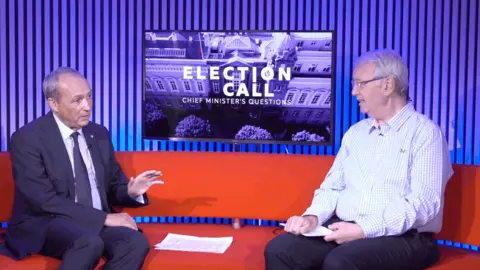 Two men sitting on a red sofa. On the left, the man has short grey hair. He is wearing a dark suit, dark tie and light shirt. On the right, the man has short grey hair and glasses. He is wearing a light blue and white checked shirt and dark trousers. Between them is a TV screen which says 'Election Call Chief Minister's Questions'.
