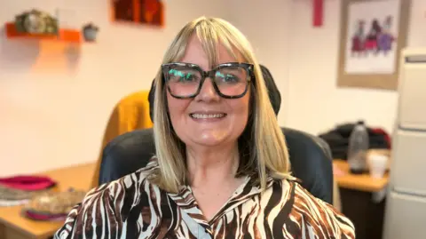 Julie Blagbrough with blond hair and wearing a brown and cream patterned shirt and tortoise shell glasses. She is in an office and smiling.