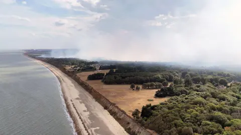 Una imagen aérea de PA Media muestra el mar a la izquierda del encuadre, con árboles, brezales y una gran columna de humo.