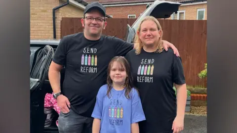 Family handout Rosie, in the front and centre frame, with her dad James and mum Julie standing behind her. They are all standing closely together, and James has his arm around Julie's shoulders. They are smiling and standing outside, in front of a dark coloured car that has a rear door and boot open. A garden fence and housing can be seen in the background. Rosie is wearing a purple T-shirt with "SEND Reform" written on it. James and Julie are wearing black T-shirts with the same slogan written on them. 