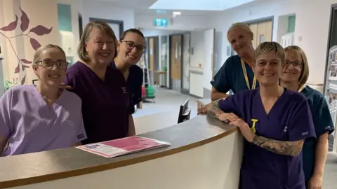 Six women in purple and blue scrubs, standing at the front desk of the hospice, smiling at the camera.