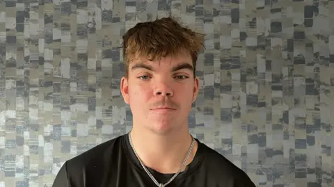 The picture shows a man wearing a black t-shirt with a silver chain around his neck. The man is standing in front of patterned wallpaper