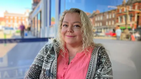 A women with long curly blonde hair, wearing a pink shirt and a patterned cardigan sat in leek town centre.