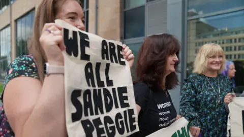 A woman holds a sign saying WE ARE ALL SANDIE PEGGIE while standing outside. Two other women are with her, also holding signs and banners.