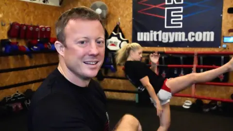 John Fairhall/BBC Richard Gent is pictured smiling to camera while wearing a black t-shirt. Turner can be seen training in the background, working on some of her kicks.